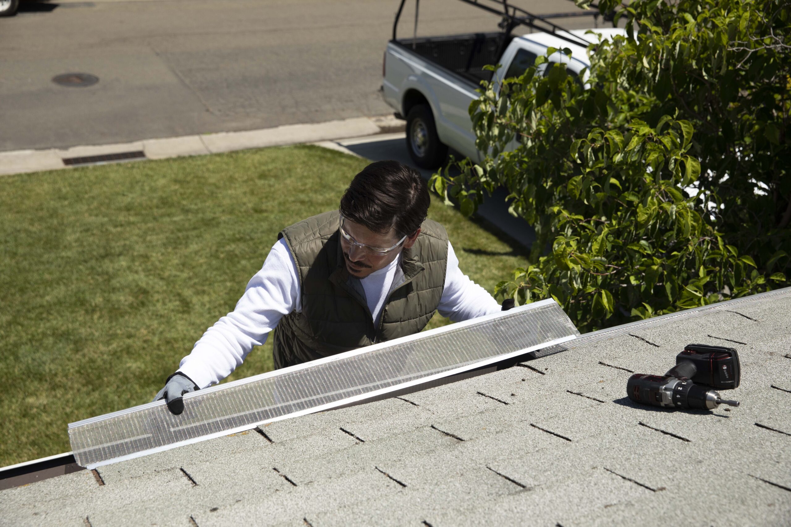 A man wearing safety glasses and gloves installs an Atlas Gutter Guard with RiserStep Technology on a house roof. A cordless drill and screws are nearby, with a white pickup truck and trees in the background.
